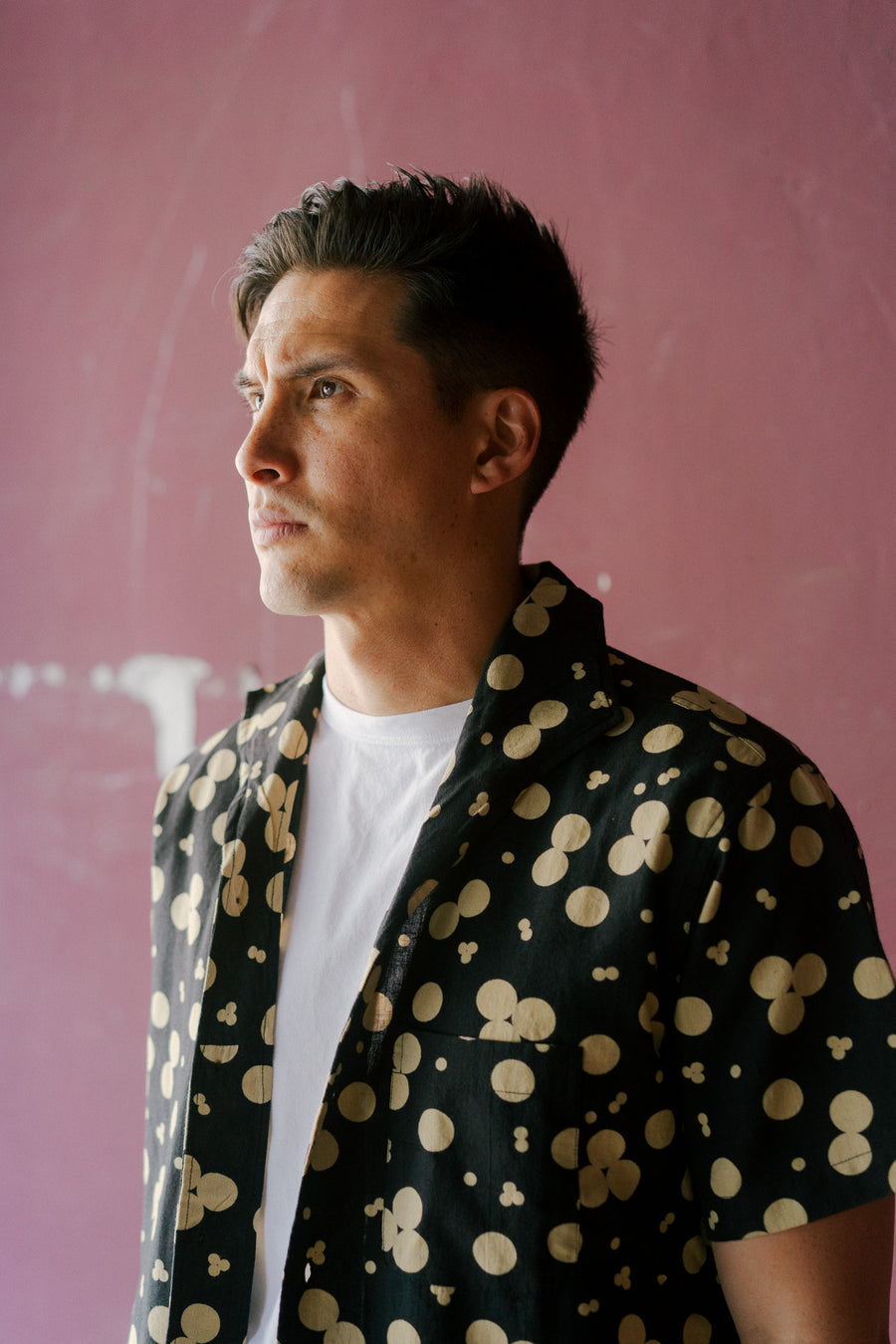 A man wears the black shirt with tan polka dots and stands against a red wall.
