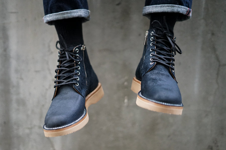 Two feet dangle freely while encased in Viberg Service Boots in a waxy navy roughout leather.