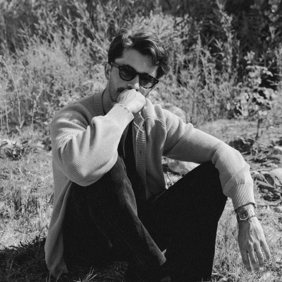 A man in sunglasses and a sweater sits on grass.