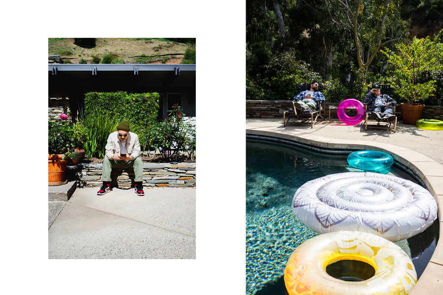A man sits on a stone wall checking his phone, while on the left two men lay in lounge chairs next to a pool.