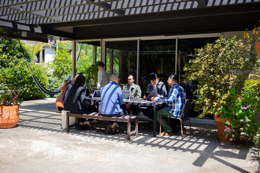 The 3sixteen team sits around a table seated on benches, intently discussing important company matters.