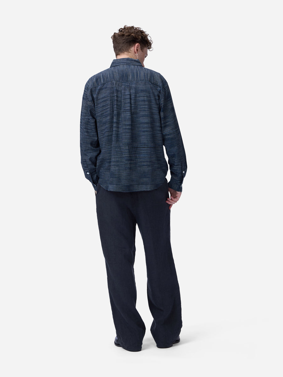 A man viewed from behind wearing a dark blue patterned button-up shirt and dark navy trousers, standing against a light gray studio background.