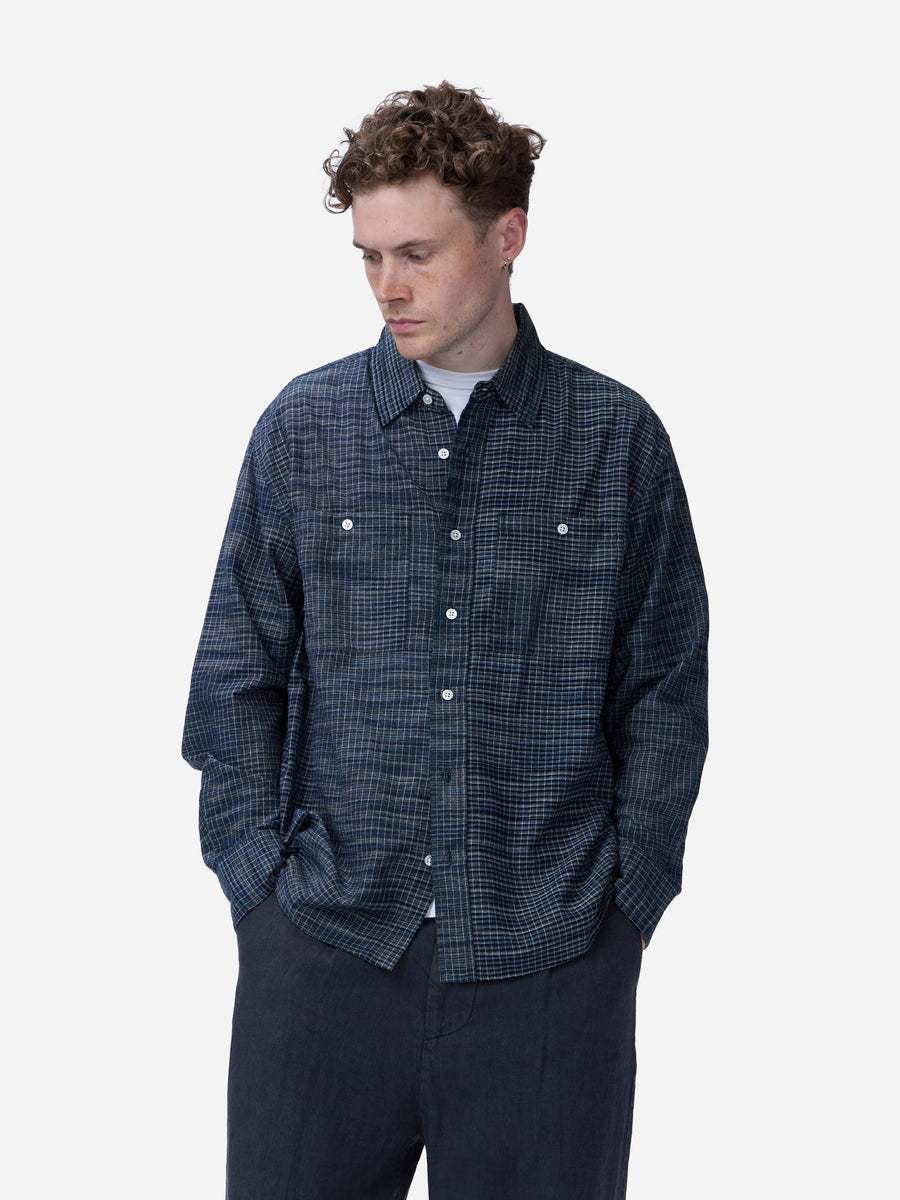 A man wearing a dark navy blue and black checkered long-sleeve button-up shirt, paired with dark trousers, photographed against a solid light gray background.