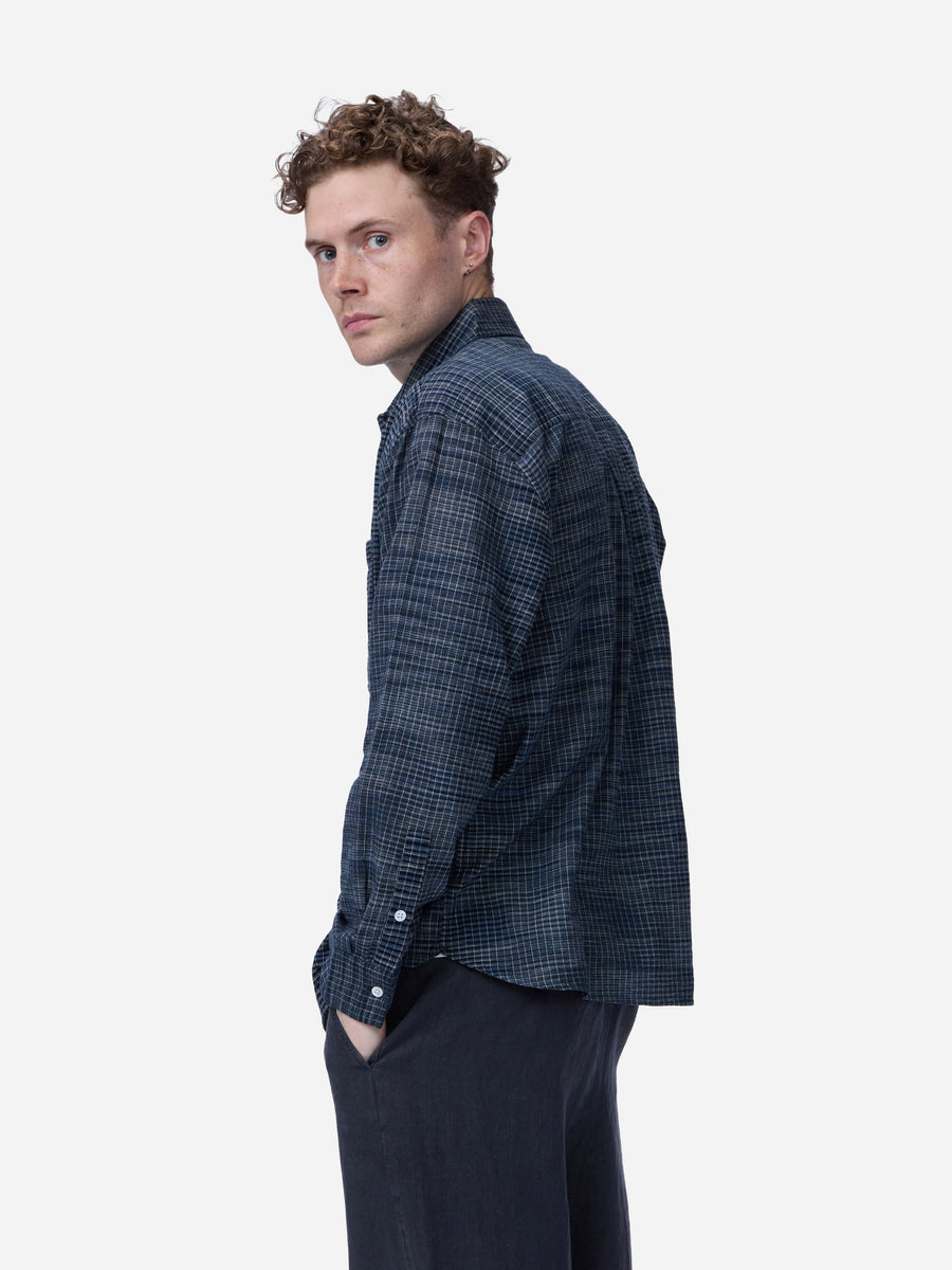 A man wearing a dark navy blue and black checkered long-sleeve shirt, shown from a side profile against a light gray background.