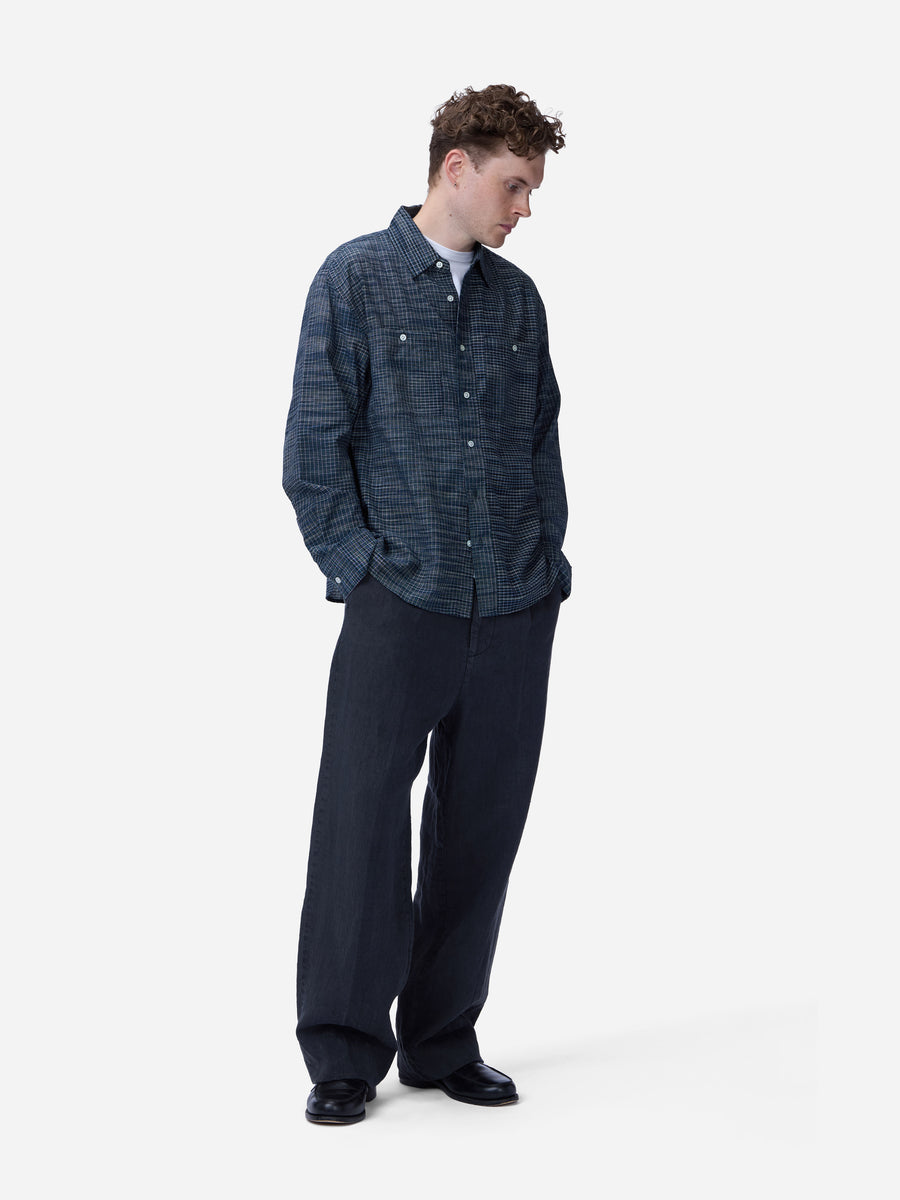 A man wearing a dark blue textured button-up shirt and dark navy wide-leg trousers, standing against a light gray studio background.