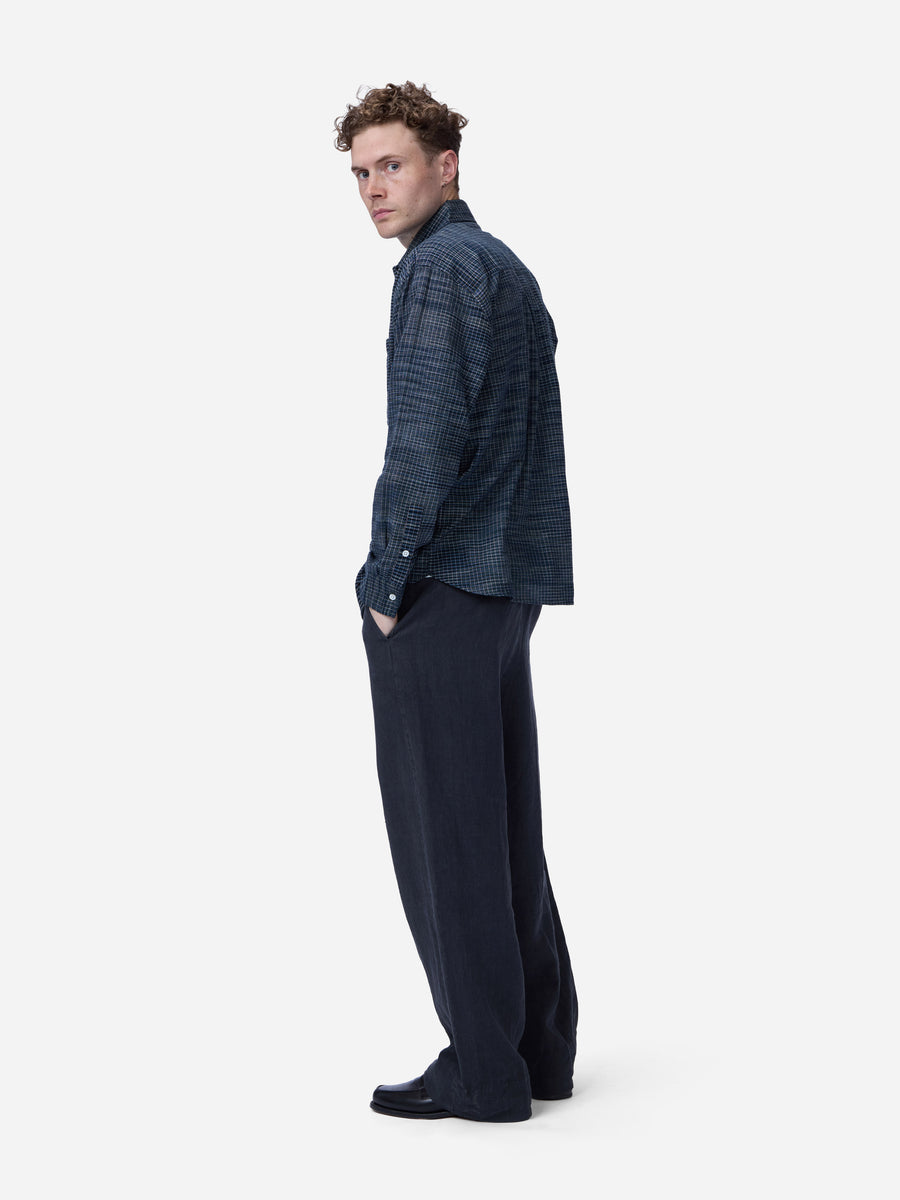 A man in a dark blue patterned shirt and dark trousers, shown from a side profile against a light gray background.