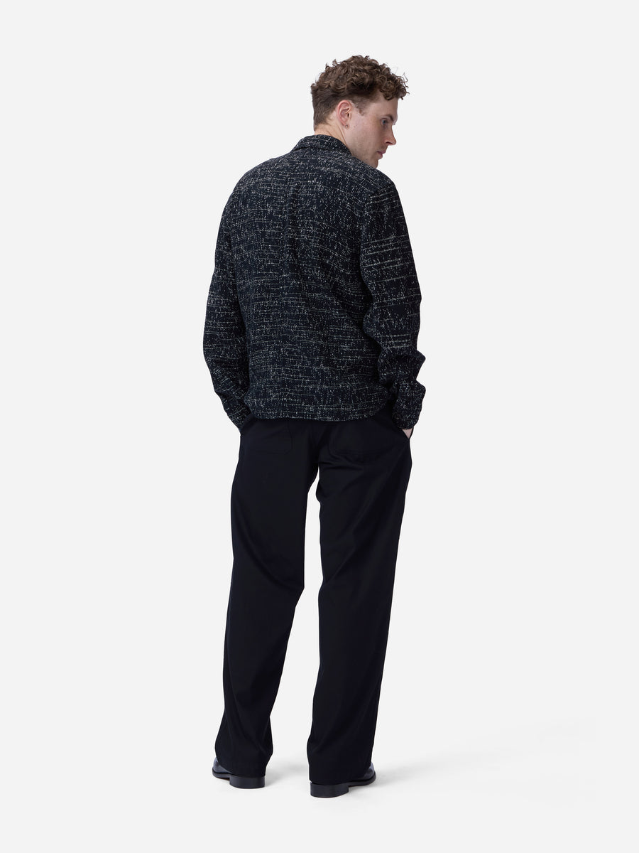 A man viewed from the back wearing a dark textured knit jacket and black trousers, standing against a plain light gray studio background.