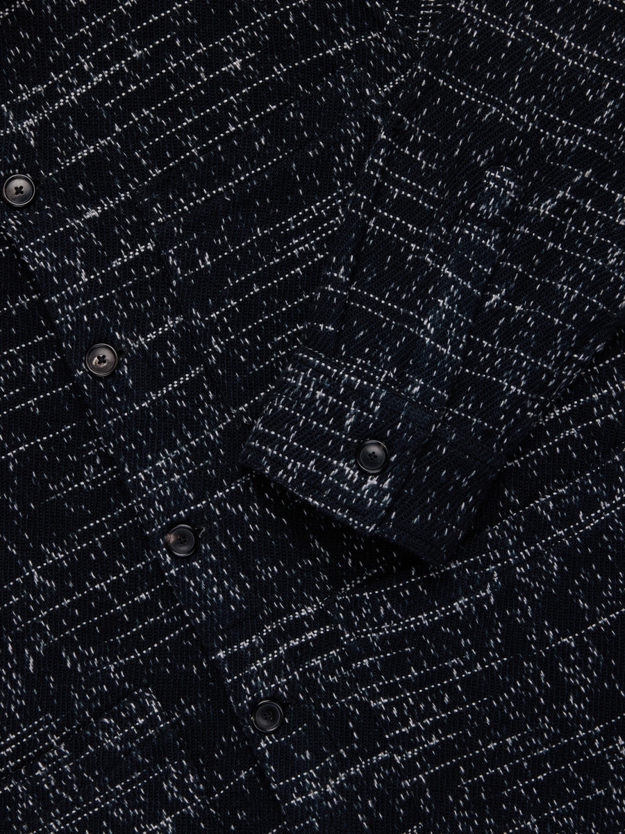 Close-up of a dark navy and black textured knit fabric with a subtle diagonal speckled pattern and dark buttons.