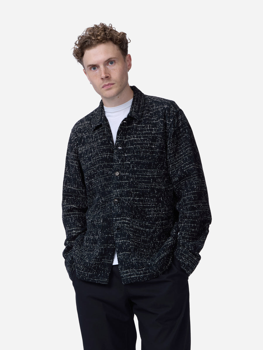 A man wearing a black and white textured tweed button-up shirt jacket over a white t-shirt and black trousers, photographed against a solid light gray background.