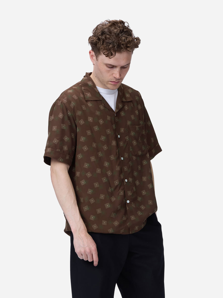 A man wearing a brown short-sleeve button-up shirt with a repeating beige geometric monogram pattern, styled over a white t-shirt and black trousers, photographed against a light gray background.