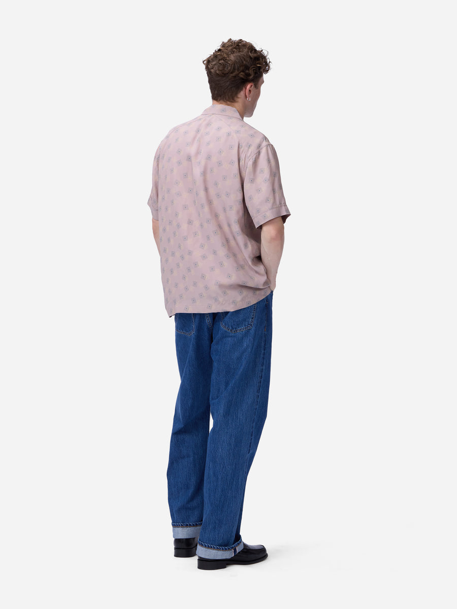 A man viewed from behind wearing a light pink short-sleeve button-up shirt with a small repeating pattern, blue jeans, and black shoes, standing against a plain light gray background.