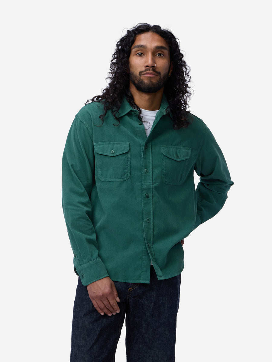 A man with long curly dark hair wearing a dark green corduroy button-up shirt over a white undershirt and dark blue jeans, standing against a light gray studio background.