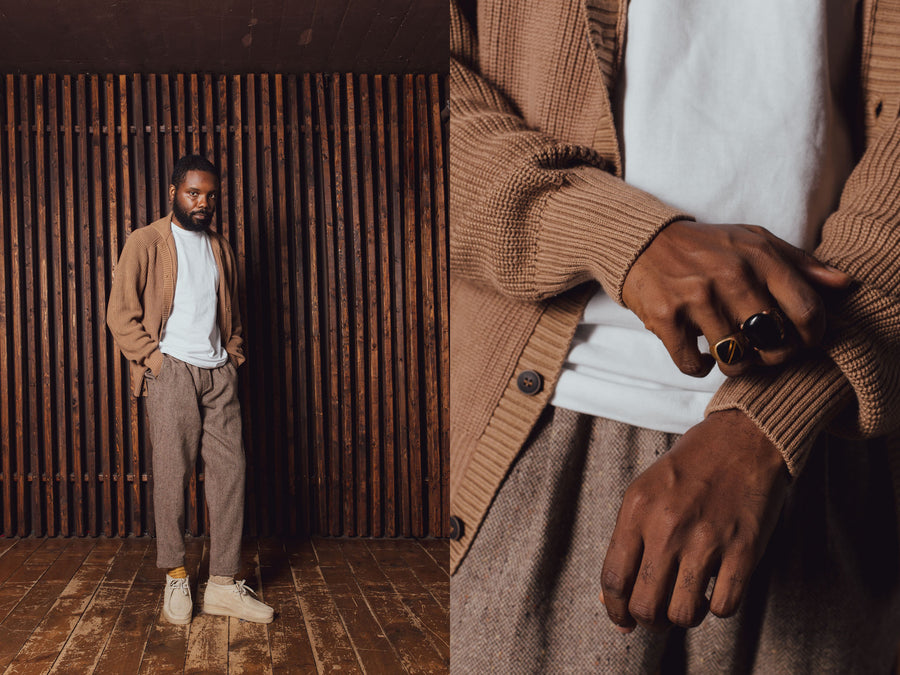 A man wears a tan sweater, white tee and wool pants.