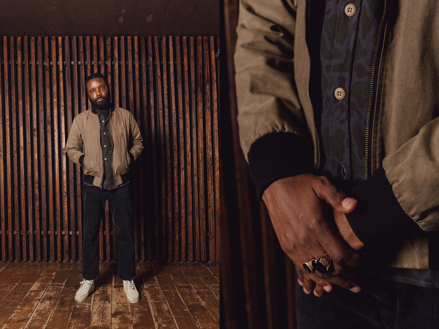 A man in black jeans and a tan bomber jacket stands in front of a wooden slat wall.