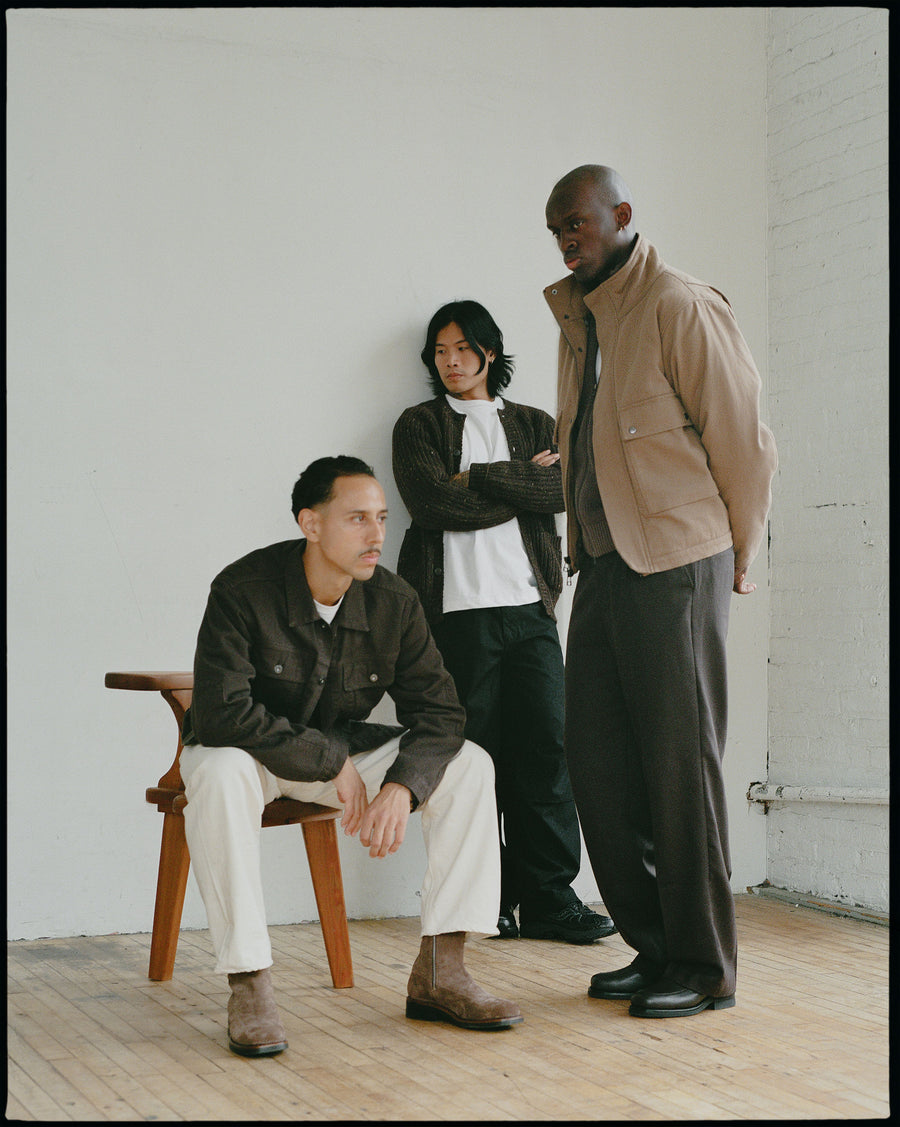 A trio of guys in earth toned clothing stand in a white room.