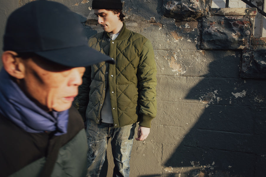 A model wears an olive down jacket while a passerby crosses in front of him.