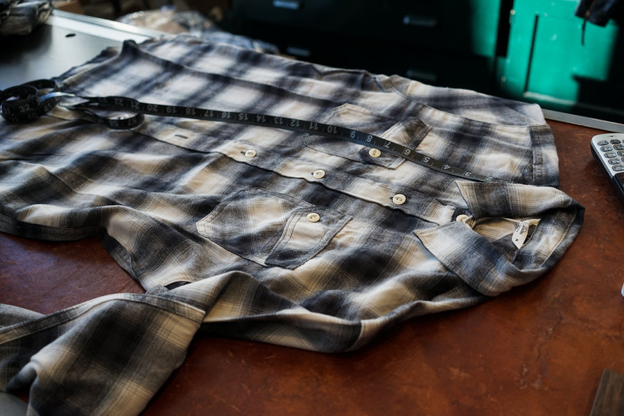 A 3sixteen shirt sits on a table with a measuring tape, where the staff is presumably taking measurements of it.