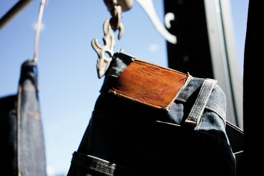 A well-worn 3sixteen jean hangs from a metal hanger, showing the darkened natural leather patch.