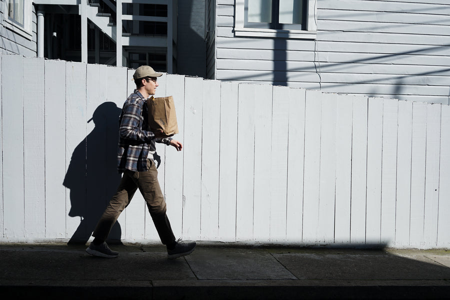 A man walks down a street carrying a brown paper bag full of sandwiches.