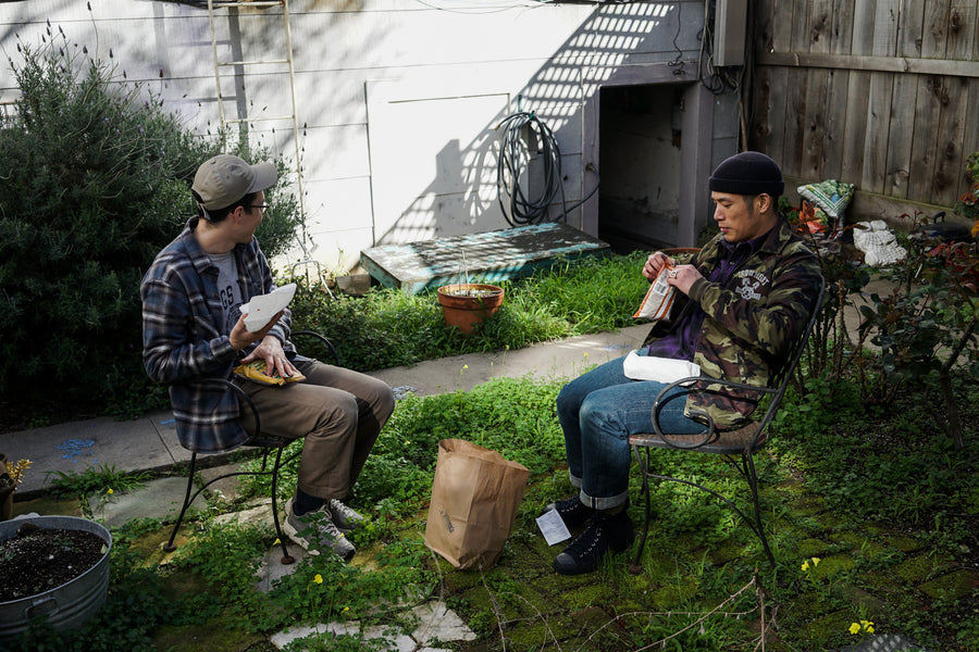 Two men sit in a backyard eating said sandwiches.