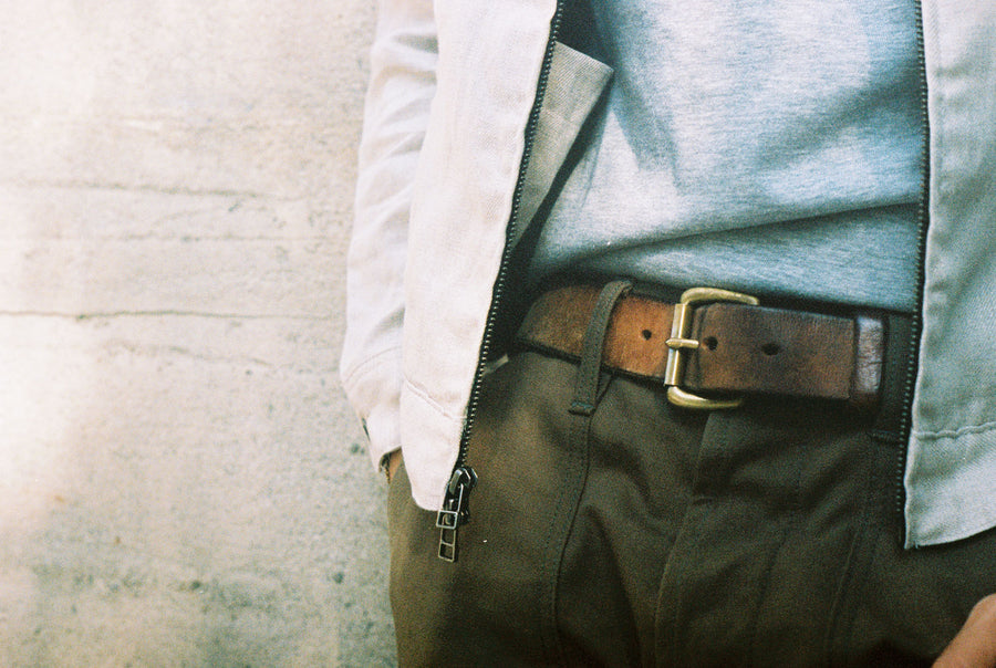 The model wears a heather grey t-shirt under the jacket, tucked into a pair of olive fatigue pants that are held up by a dark tan leather belt.