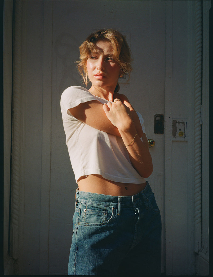 A lady in a cropped white tee and light blue jeans stands while the sun sets.