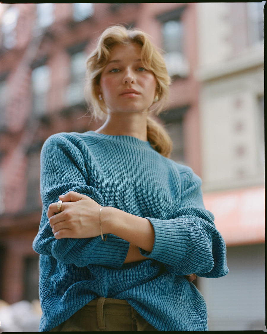 A lady in a blue sweater and tan pants stands outside. 