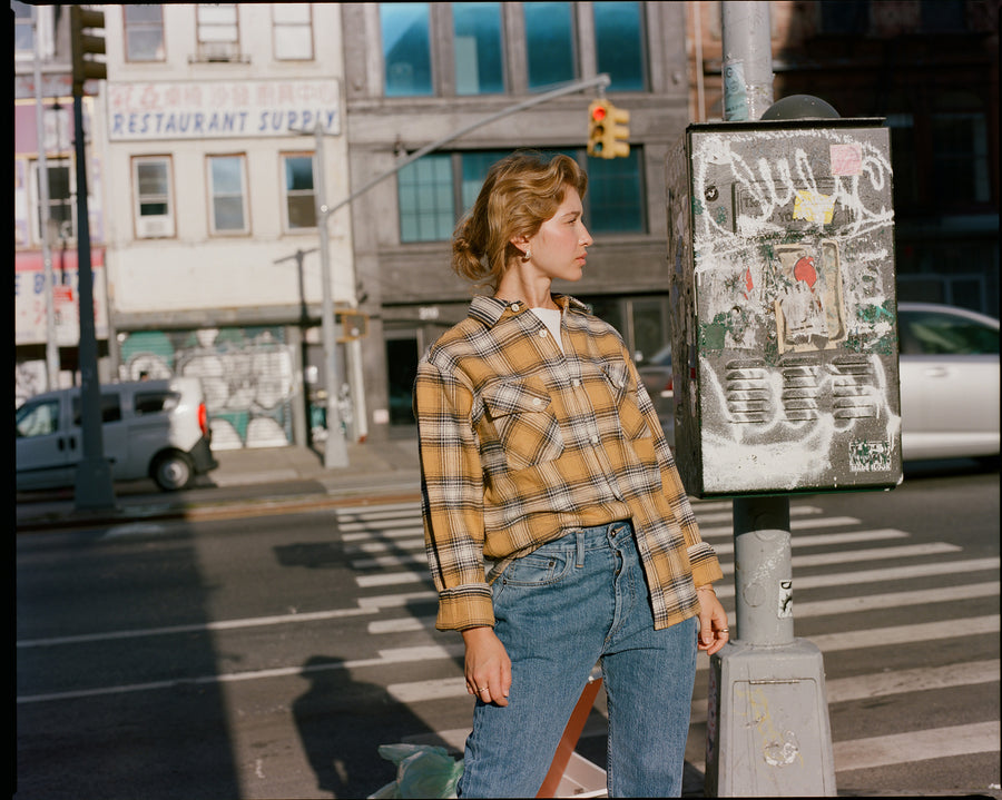 A lady in a yellow flannel and blue jeans stands on a street.