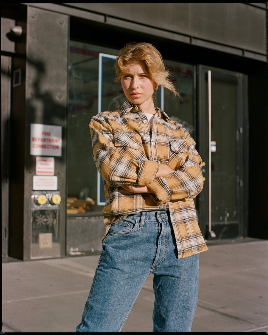 A lady in a yellow flannel and light blue jeans stands in the sunlight on a sidewalk.