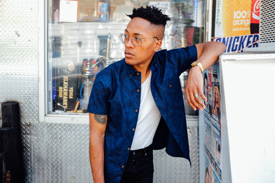A man wears the indigo bandana short sleeve shirt open over a white tank top while looking right.